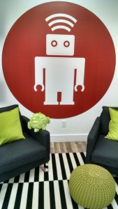 thoughtbot robot logo painted on the wall, with a poofy android robot sitting in front of it